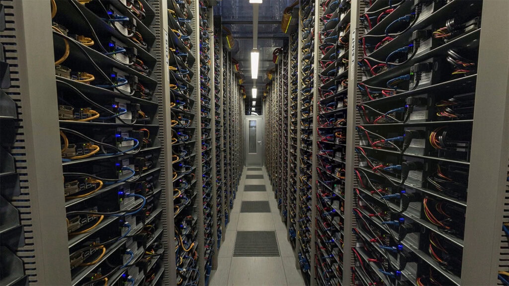 A screenshot from the Hetzner 360° tour. We're in a narrow corridor inside a server farm, surrounding by shelves filled from floor to ceiling with server equipment and cables.